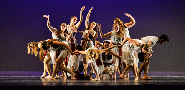 a group of dancers wearing loosely draped white cloth, dispersing from a circle on stage in front of a purple and black backdrop