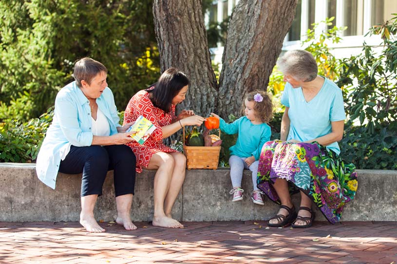 Education teachers sit with a toddler along the library wall under a tree