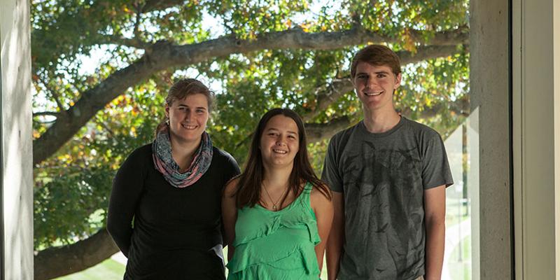 Bryn Athyn College students stand together smiling in the student lounge in front of a window with an oak tree in the background.