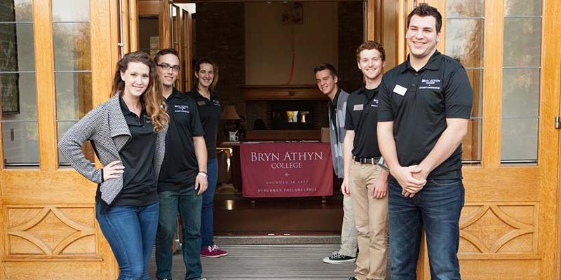 Student Ambassadors greet the camera outside the Brickman Great Hall front doors at Bryn Athyn College